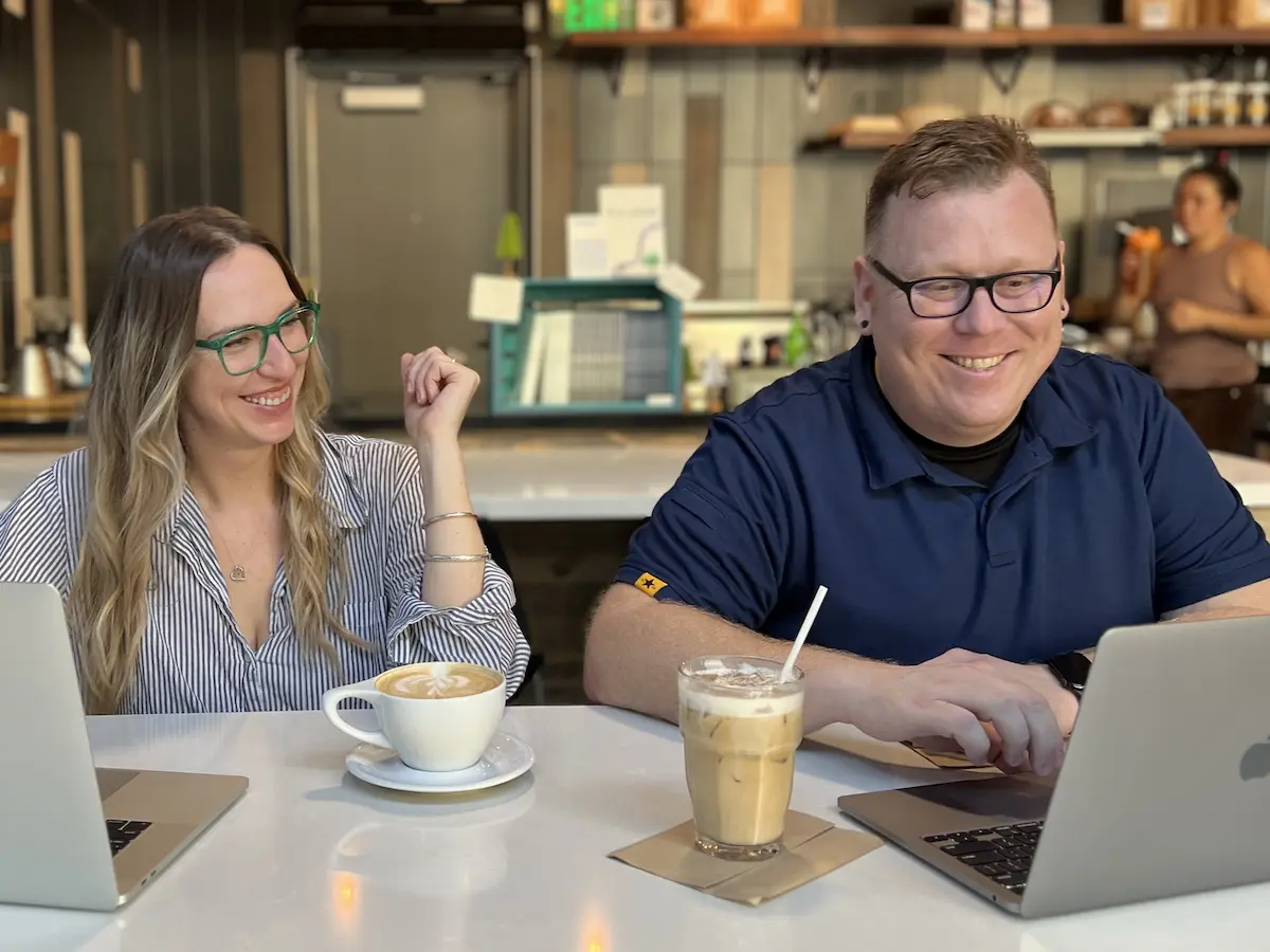 PSI Seminars Podcast hosts Tim and Mackenzie smiling while working on laptops at a coffee shop table