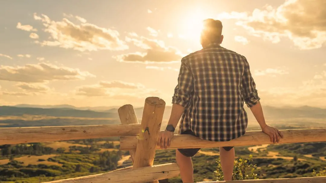 Person sitting on a wooden fence overlooking a sunlit mountain landscape at sunset, reflecting in a peaceful setting